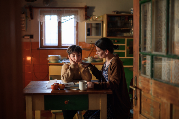 A poor mature mother and sad small daughter learning indoors at home, poverty concept.