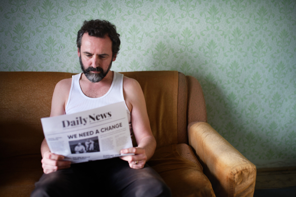 Front view of poor mature man reading newspapers indoors at home, poverty concept.