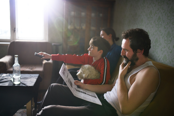 Portrait of poor mature couple with small daughter watching TV indoors on sofa at home, poverty concept.