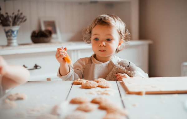 A happy small toddler girl sitting at the table, decorating cakes at home.