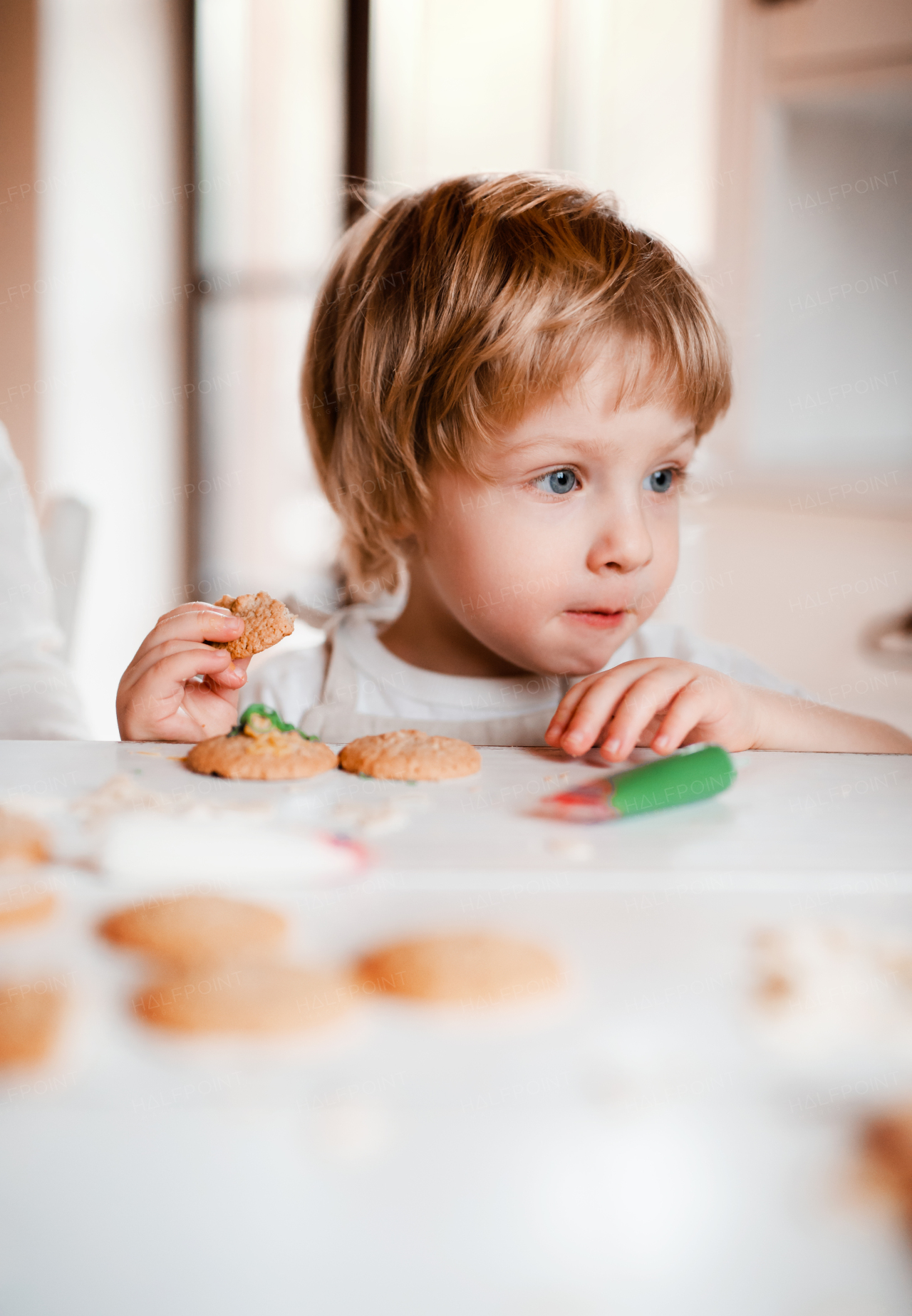 A small happy toddler boy sitting at the table, decorating and eating cakes at home.