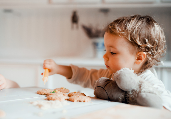 A happy small toddler girl sitting at the table, decorating cakes at home.