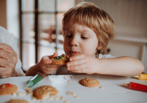 A small happy toddler boy sitting at the table, decorating and eating cakes at home.