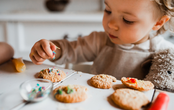 A happy small toddler girl sitting at the table, decorating cakes at home.