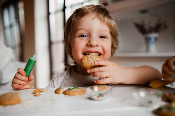 A small happy toddler boy sitting at the table, decorating and eating cakes at home.