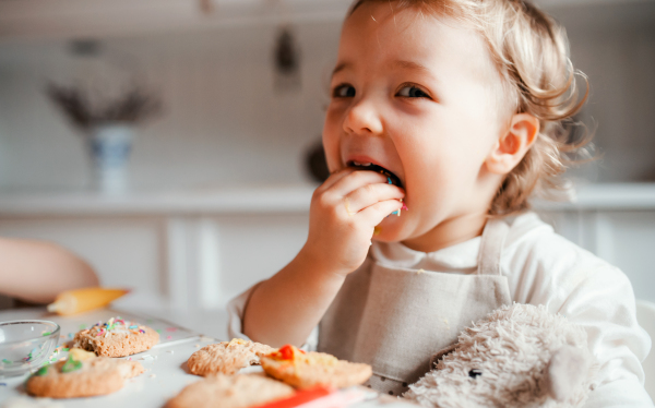 A happy small toddler girl sitting at the table, decorating cakes at home.