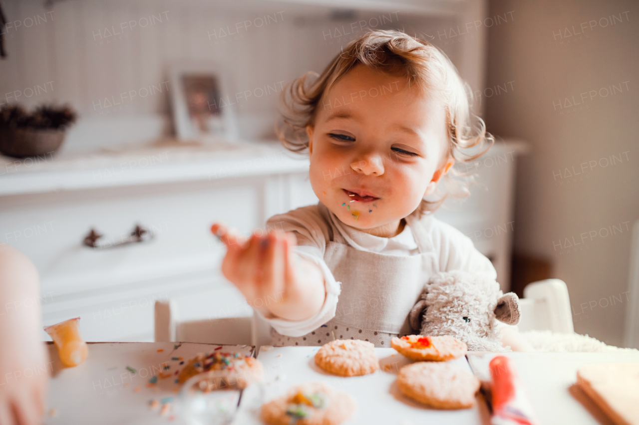 A happy small toddler girl sitting at the table, decorating cakes at home.