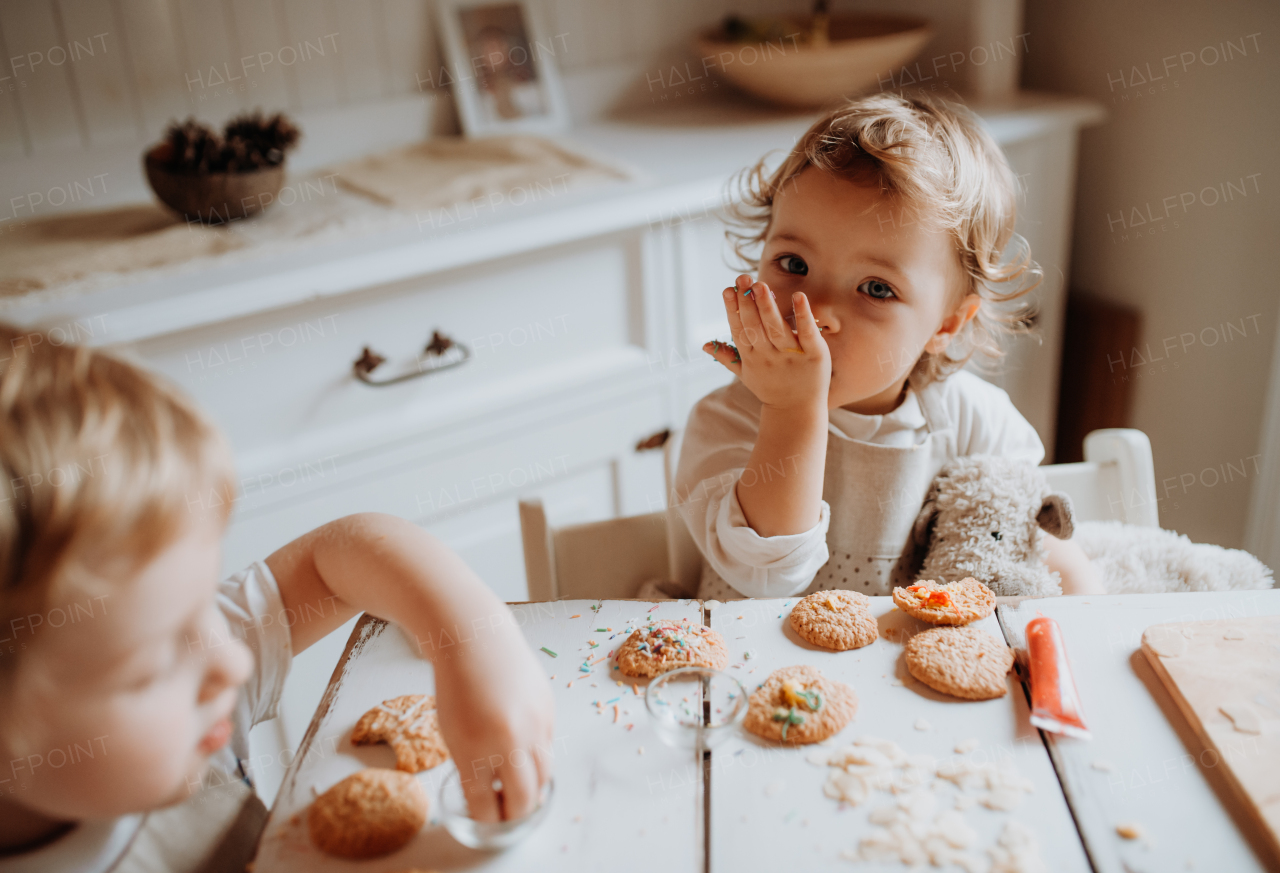 Two happy small toddler children sitting at the table, decorating and eating cakes at home.