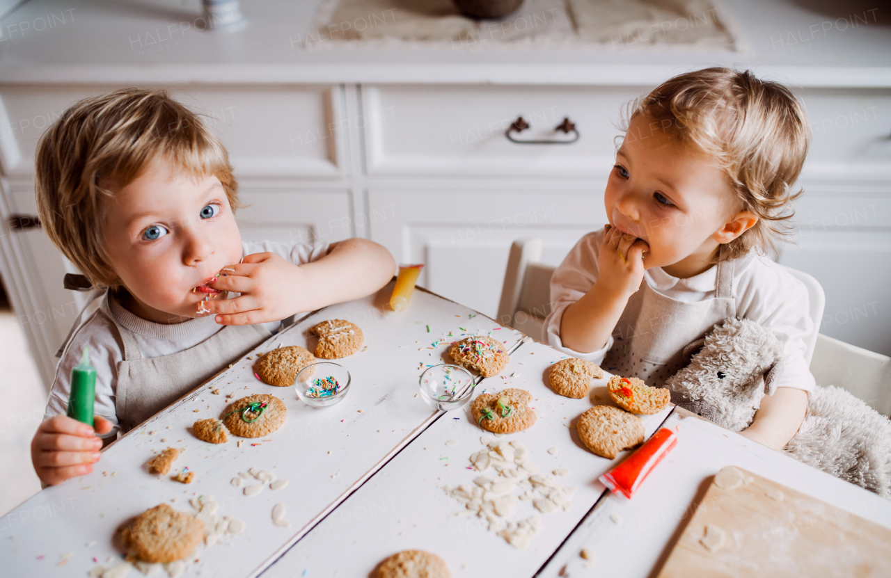 Two happy small toddler children sitting at the table, decorating and eating cakes at home.