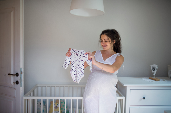 Front view portrait of pregnant woman indoors at home, holding girls bodysuit.