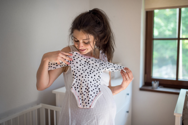 Front view portrait of pregnant woman indoors at home, holding girls bodysuit.