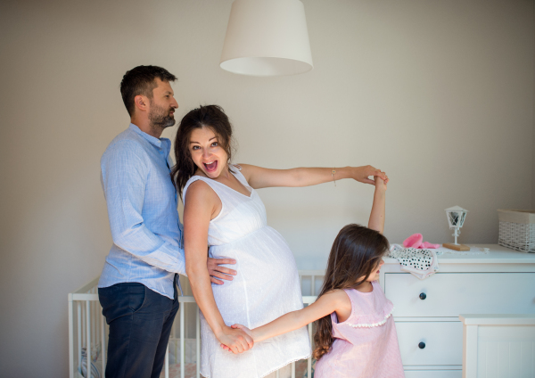 Side view portrait of cheerful pregnant woman with husband and small daughter indoors at home.