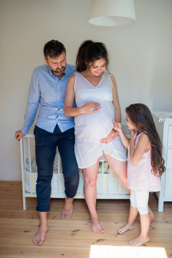 Front view portrait of pregnant woman with husband and small daughter indoors at home.