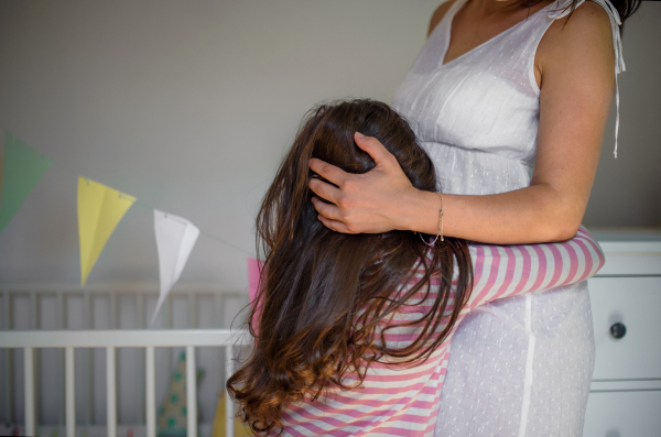 Portrait of unrecognizable pregnant woman with small daughter indoors at home, hugging.