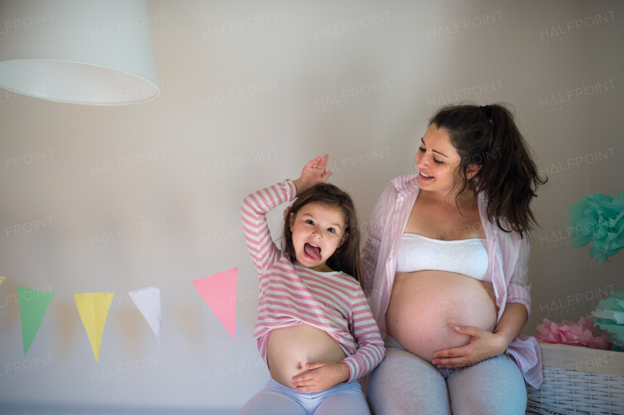 Portrait of happy pregnant woman with small daughter indoors at home, showing belly.