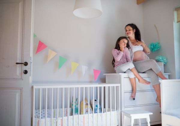 Portrait of happy pregnant woman with small daughter indoors at home, sitting on chest of drawers.