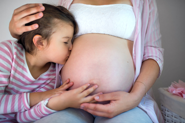 Side view of small girl kissing belly of unrecognizable pregnant mother indoors at home.