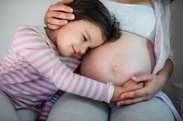 Portrait of unrecognizable pregnant woman with small daughter indoors at home, hugging.