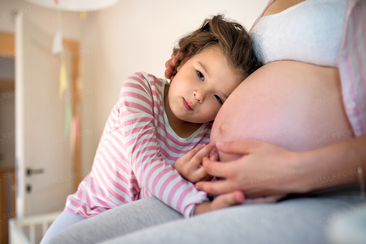 Portrait of small girl hugging belly of unrecognizable pregnant mother indoors at home.