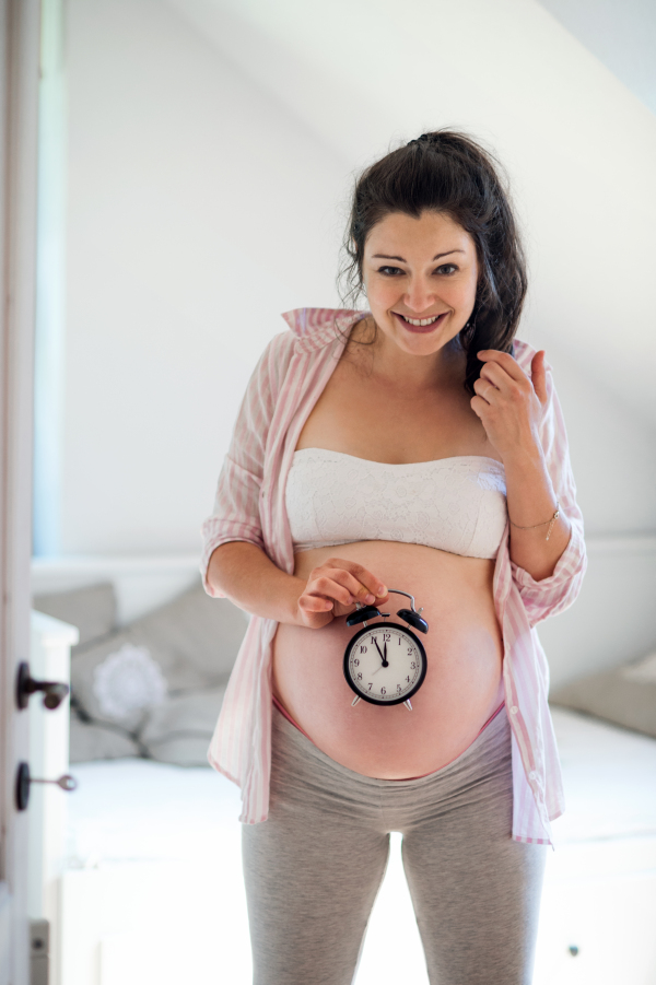 Portrait of happy pregnant woman with clock indoors at home, due date concept.