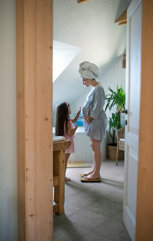 Side view portrait of pregnant woman with small daughter indoors in bathroom at home.