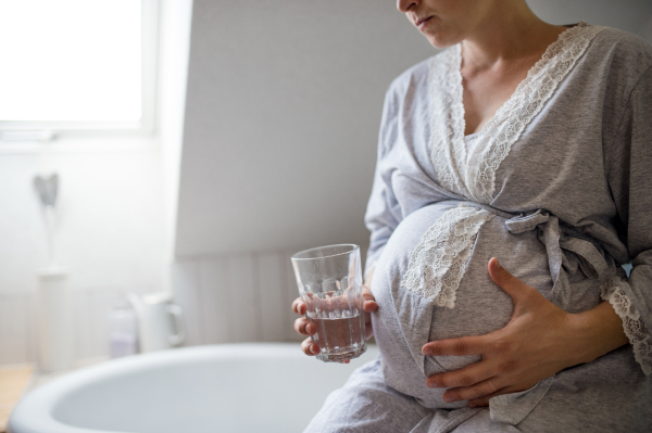 Midsection of unrecognizable pregnant woman indoors in bathroom at home, holding glass of water.