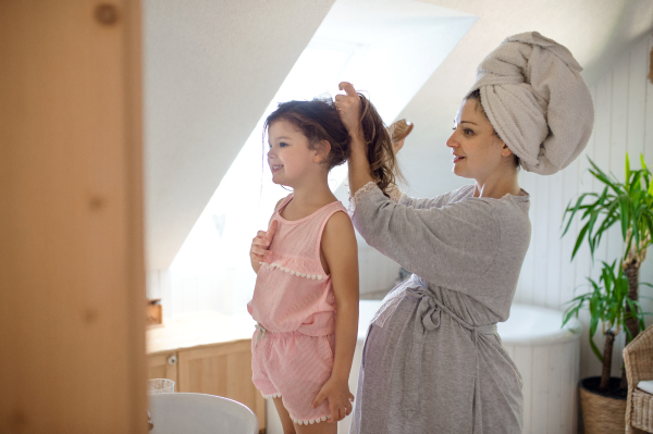 Portrait of pregnant woman with small daughter indoors in bathroom at home, combing hair.