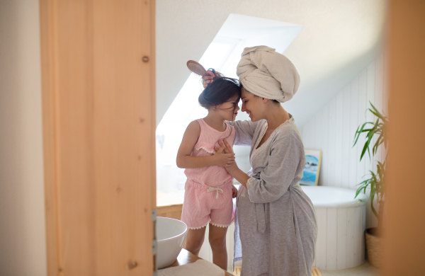 Side view portrait of pregnant woman with small daughter indoors in bathroom at home, brushing hair.