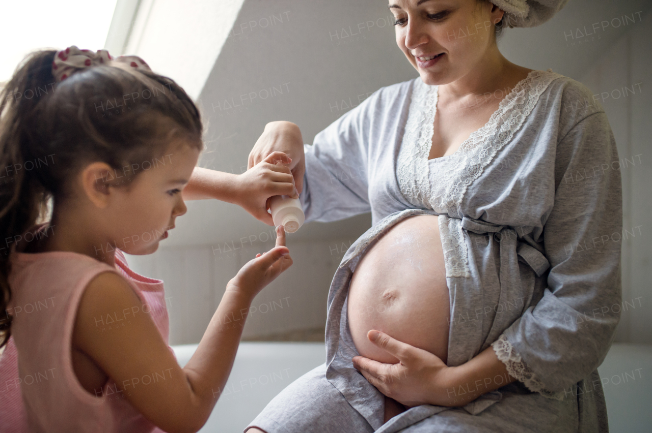 Happy small girl creaming belly of pregnant mother indoors in bathroom at home.