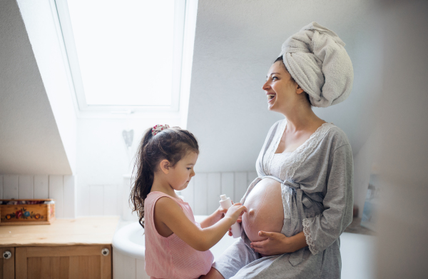 Happy small girl creaming belly of pregnant mother indoors in bathroom at home.