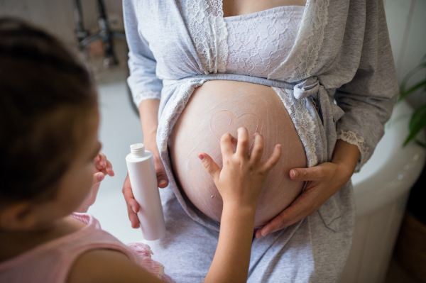 Unrecognizable small girl creaming belly of pregnant mother indoors in bathroom at home.