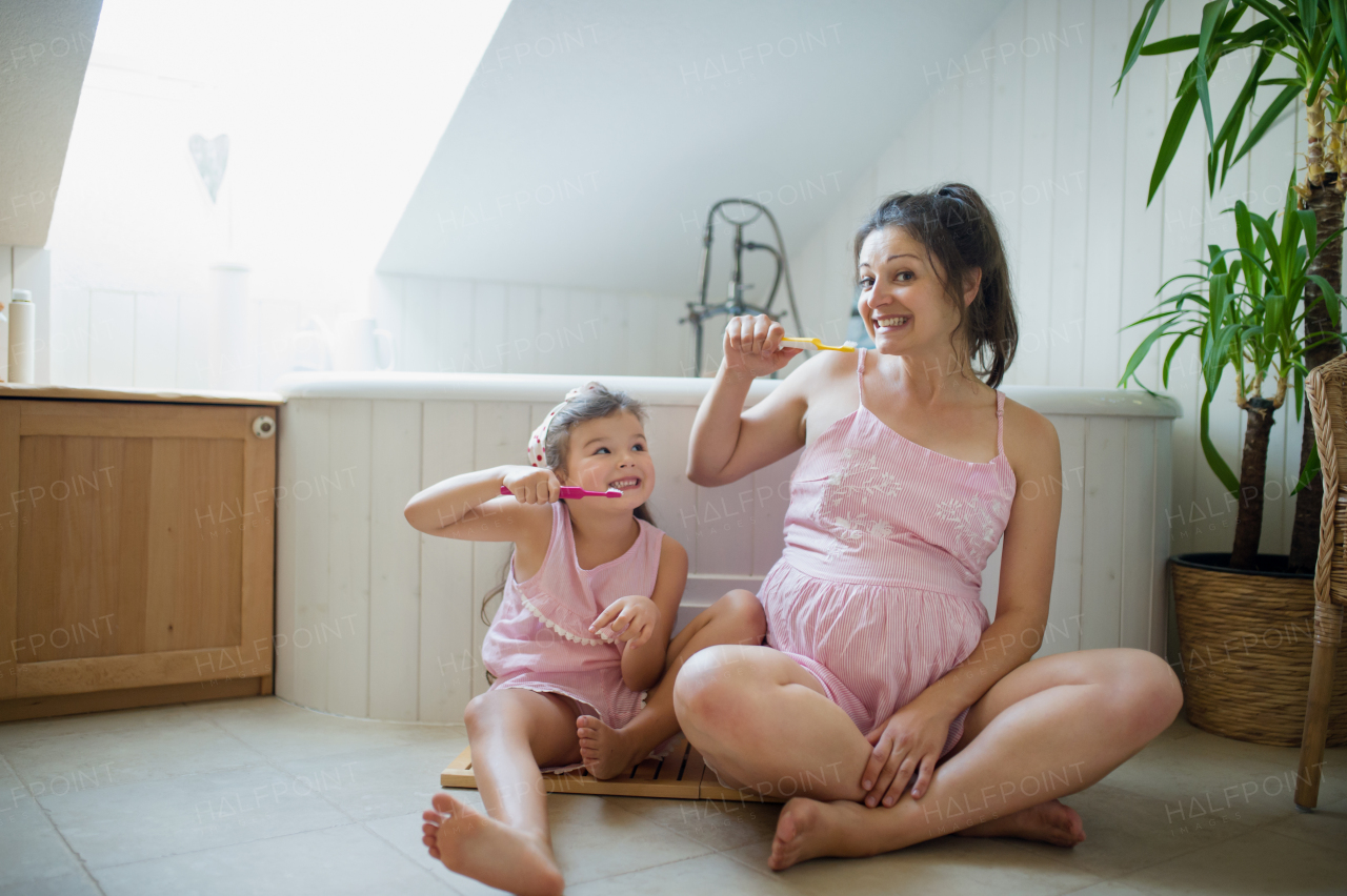 Portrait of happy pregnant woman with small daughter indoors in bathroom at home, brushing teeth.