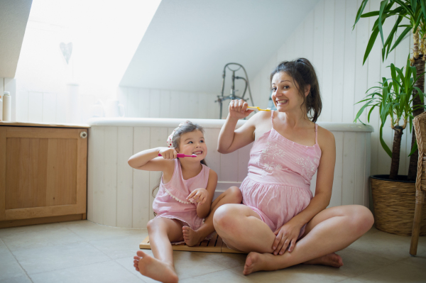 Portrait of happy pregnant woman with small daughter indoors in bathroom at home, brushing teeth.