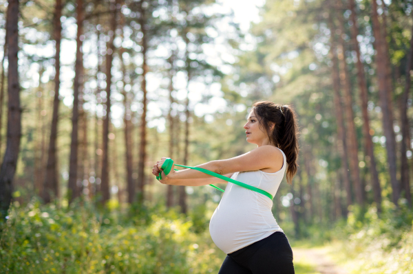 Side view portrait of happy pregnant woman outdoors in nature, doing exercise with elastic band.