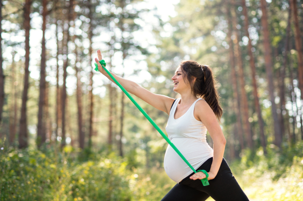 Side view portrait of happy pregnant woman outdoors in nature, doing exercise with elastic band.