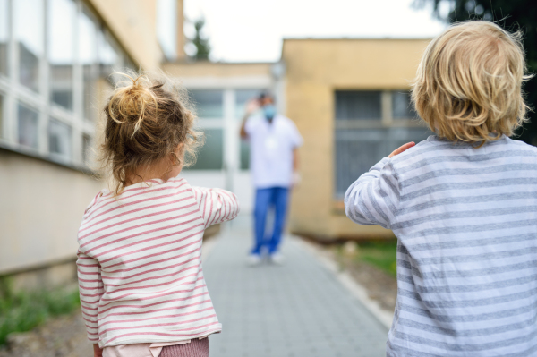 Rear view of children waving at father doctor in front of hospital, coronovirus and isolation concept.