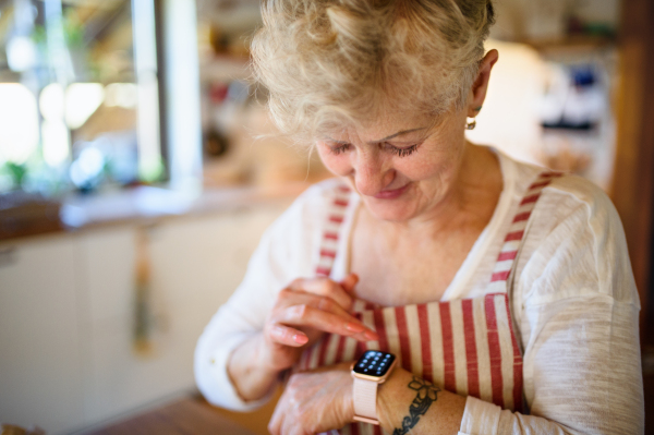 A senior woman with apron indoors at home, using smartwatch.