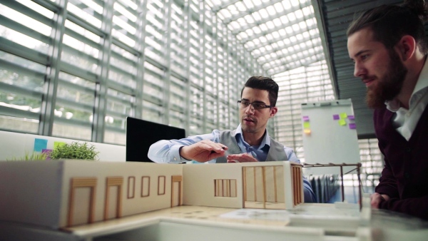 Two young male architects with model of a house in office, working and talking. Slow motion.