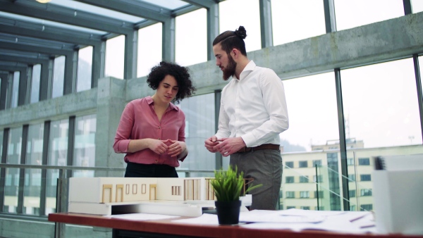 Two young architects with model of a house standing in office, working and talking.