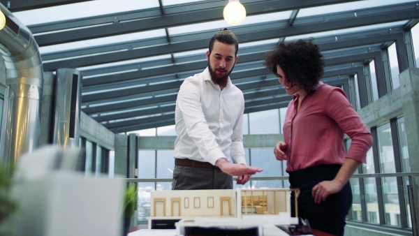 Two young architects with model of a house standing in office, working and talking.