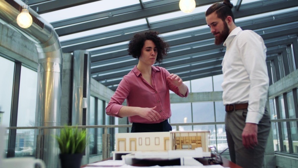 Two young architects with model of a house standing in office, working and talking.