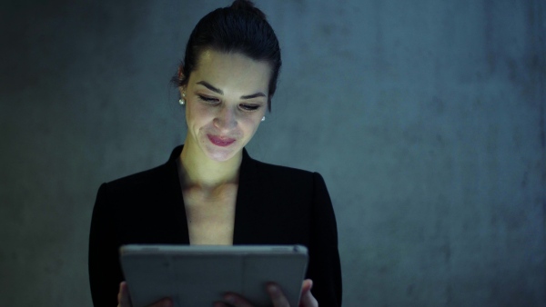 A portrait of happy young business woman with digital tablet standing against concrete wall in office.