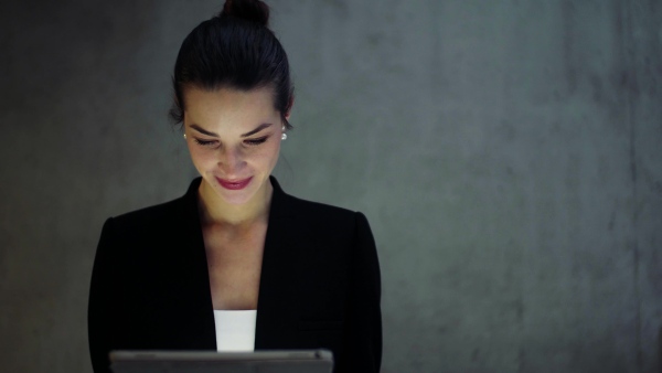 A young business woman with tablet walking in office, concrete wall in background.