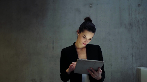 A young business woman with tablet walking in office, concrete wall in background.