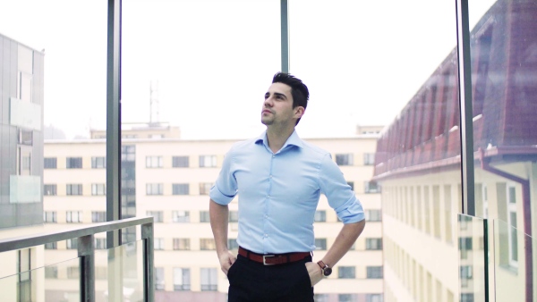 A portrait of young businessman walking indoors in an office, hands in pockets.