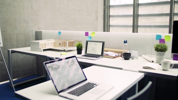 Laptops, blueprints on screens and model of a house in interior of office of architects.