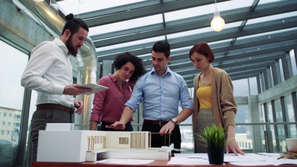 A group of young architects with model of a house standing in office, working and talking.