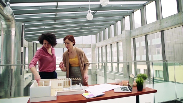 Two female young architects with model of a house standing in office, working and talking. Slow motion.