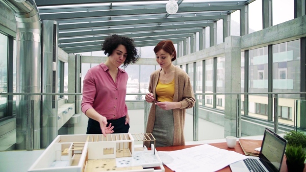 Two female young architects with model of a house standing in office, working and talking. Slow motion.