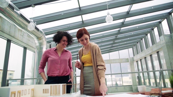 Two female young architects with model of a house standing in office, working and talking. Slow motion.